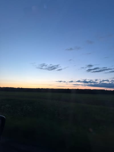 The image depicts a serene rural landscape at twilight or sunset, seen from a perspective that suggests viewing from a vehicle. The sky dominates the upper two-thirds of the frame, transitioning from a deep indigo blue at the top to a lighter, softer blue, and finally to a warm band of golden yellow and faint orange light hugging the horizon. Scattered, wispy clouds, dark against the fading light, are visible across the blue expanse.
A distinct, dark silhouette of a forest or dense tree line marks the horizon, stretching horizontally across the width of the image. Below this dark line, the landscape consists of fields, appearing dark green to nearly black due to the low light, with subtle textures suggesting agricultural patterns. In the very bottom right corner of the image, a dark, indistinct shape, possibly part of a vehicle's dashboard or window frame, is visible, indicating the photograph was likely taken from inside a car or similar transport.
There are no people or visible text in the image. The scene captures the peaceful transition from day to night over the natural environment of Yus'ki, Russia, characterized by vast skies and expansive land.