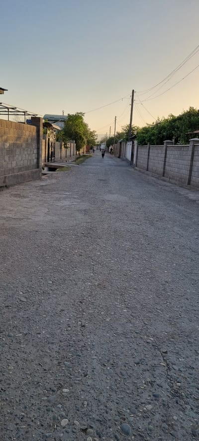 The image shows a residential street in the city of Khovaskor, Tajikistan. The street is paved with gravel and is lined with brick walls on both sides. There are houses and trees visible behind the walls. In the distance, two people are walking down the street, and a motorcycle is also visible. The sky is clear and blue, with a hint of sunset colors on the horizon. The scene appears to be quiet and peaceful.
