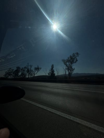A car interior shows a sunlit sky. A road stretches to the side, lined with sparse trees. The sun glares brightly in the sky, casting long rays across the scene. A hint of distant mountains lies near the road. A dark object, possibly part of the vehicle, intrudes at the bottom, partially obscuring the view. The overall mood is bright and somewhat obscured by the sun's intensity.