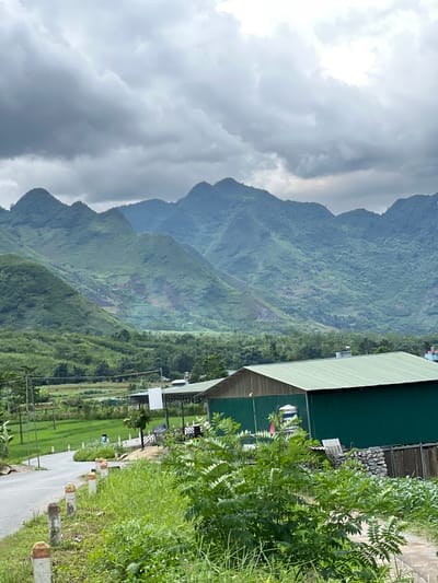The image depicts a rural landscape in Lùng Tám Commune, Vietnam, dominated by lush green mountains under a cloudy sky. In the foreground, a paved road curves through a grassy area with roadside markers. A low-lying green building with a corrugated metal roof is visible on the right, partially obscured by dense green foliage. Beyond the building, a stone wall and wooden fencing are present. In the middle ground, a person is walking along the road in front of a field. The background is filled with rolling, verdant mountains, some showing signs of erosion or cultivation. The overall atmosphere is overcast and serene, characteristic of a rural Vietnamese setting.
