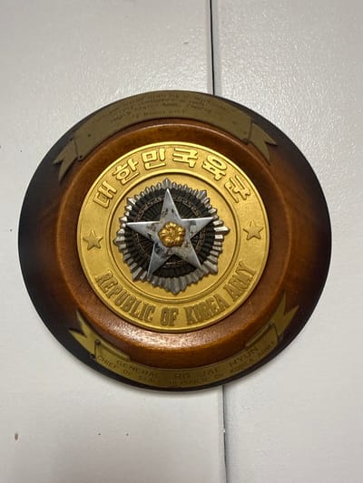 A round, wooden plaque features a gold-colored emblem with Korean writing and the words "REPUBLIC OF KOREA ARMY" in English lettering. A silver, star-shaped device with a gold floral symbol rests in the emblem's center. Two ribbons flank the emblem on the plaque's edge, bearing printed text. One ribbon mentions "WILLIAM" and "MARCH 16." The opposite ribbon references "RO JAE HYUN," "GENERAL," and "CHIEF OF STAFF, REPUBLIC OF KOREA ARMY."