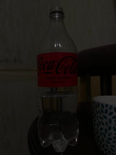 A partially full bottle of Coca-Cola Zero Sugar is positioned vertically in the center of the frame, against a dark, textured background. The bottle has a red label with the Coca-Cola logo in black script. Below the logo, the words "ZERO SUGAR" are visible in black text, followed by smaller text that appears to be in a different language. The bottle is made of clear plastic and contains a clear liquid, with the liquid level reaching about half way up the bottle. The bottom of the bottle shows the typical molded plastic design. To the right of the bottle, a portion of a white mug with a blue leaf-like pattern is visible. The scene is dimly lit, with shadows obscuring much of the environment. No people are present in the image. The location of Dolakovo, Russia is not discernible from the visual information provided.