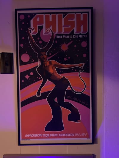 A vertically oriented Phish New Year's Eve 98/99 concert poster is centrally depicted, mounted on a light-colored wall within an interior room. The poster features a stylized humanoid figure with large, upward-curving horns and a pointed tail, rendered as a dark silhouette. The figure is set against a cosmic background featuring pink and orange planets, stars, and swirling bands of color. Text on the poster identifies the event as "New Year's Eve 98/99" and lists the dates "DEC. 28, 29, 30, 31" with the venue "MADISON SQUARE GARDEN NY, NY." To the left of the poster, a dark, rectangular wall-mounted electrical switch or dimmer, characterized by vertical ridges, is visible. The scene is illuminated by a distinct purple ambient light, casting a glow on the wall and elements within the image. A light-colored horizontal trim is present on the wall below the poster. The exact location within the city of Winooski, United States of America, cannot be determined from the provided image.