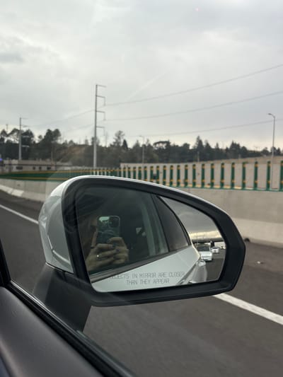 The image shows a side mirror of a white car, presumably taken from within the car. The mirror's reflection shows the person in the driver’s seat is holding a phone and taking a picture. The mirror has the warning text "OBJECTS IN MIRROR ARE CLOSER THAN THEY APPEAR" on it. Outside the car, the highway has a green and white roadside barrier and a line of trees and foliage in the distance. The sky is overcast and gray. The location is likely Turcio Primera Sección, Mexico, as it is a highway driving in a car.
