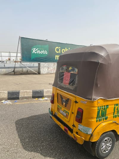 The scene depicts a static, outdoor environment in Gobirawa, Nigeria, under a bright, hazy sky. In the foreground, the rear three-quarter view of a yellow, three-wheeled passenger vehicle, commonly known as a Keke Napep, occupies the right side of the frame. The vehicle features a brown canopy top, a rear gridded panel with the text "F150" and "KEKE NAPEP" visible, two red rectangular taillights, and a partially obscured Nigerian license plate with "UGA-541YL". The rear window is a clear plastic panel, appearing worn, with colorful fabric or items visible through it. The vehicle casts a distinct, dark shadow onto the asphalt road surface.

To the left of the Keke Napep, a black and white striped curb runs parallel to the road, bordering a concrete elevated area. Scattered white and colorful plastic litter is visible on this concrete surface. Further back, a large rectangular billboard stands prominently. The billboard has a dark green background with a white "Knorr" logo (including "SINCE 1838") on the left side, and partially visible white text "Ci obi m" on the right. Behind the billboard and concrete area, indistinct structures and a hazy, light-colored sky complete the background. No individuals are present in the image.
