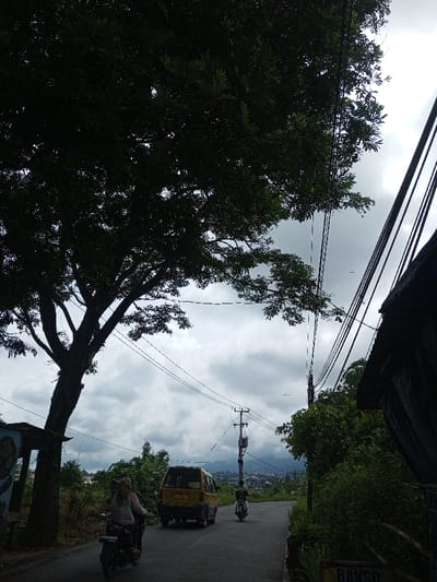A winding road leads uphill under a cloudy sky. A large tree obscures the view on the left. A yellow van and two motorcycles are on the road. Utility poles and wires line the street. Lush greenery grows on either side. A partial sign for "Bakso" is visible in the foreground. Buildings can be seen in the distance.