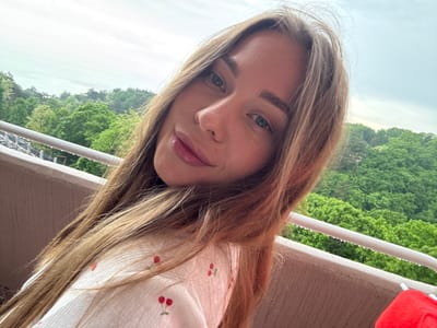 The image is a selfie of a woman with long, light brown hair, wearing a white dress with a cherry print. She is outdoors, with a background of green trees, likely on a balcony or raised platform as indicated by the white railing in the background. The location appears to be Sosnovy, Russia, based on the presence of certain architectural structures in the background. The sky is overcast. The woman is looking directly at the camera with a slight smile. A red object, potentially part of the balcony's infrastructure, is visible at the top left of the frame.