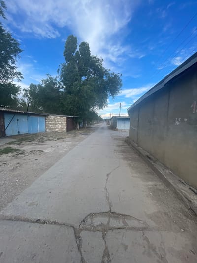 The image shows a view of a concrete alleyway lined with garages on one side and trees and sky on the other. The perspective is from a low angle, looking down the alley. On the left side of the alley, there are several garages with blue metal doors. Behind the garages, there are tall trees with green foliage, and beyond the trees, a bright blue sky with scattered white clouds. The sky and trees occupy the upper left portion of the image.
The right side of the alley is dominated by a rough, cracked concrete surface, possibly a pavement or road. The concrete has a worn and weathered appearance, with visible cracks and debris. A wall, also made of concrete, runs along the edge of the alley on this side, casting a slight shadow.
The scene appears to be set in a residential or industrial area, likely a garage complex. The time of day seems to be daytime, given the bright sunlight and clear blue sky. The weather appears to be pleasant and sunny. There are no people visible in the image, and no specific activity is taking place; it's a static scene of an outdoor environment.
The overall impression is one of a somewhat utilitarian and aged urban or suburban setting. The contrast between the natural elements of the trees and sky and the man-made structures of the garages and concrete alley is prominent. No visible text is present in the image.