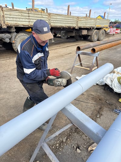 A man wearing a blue and black jacket, dark pants, red gloves, and a blue baseball cap is using an angle grinder to cut a large grey pipe. The pipe is supported by a grey metal stand. In the background, a large, rusty truck with a flatbed is parked. Several long, rusty pipes are laid out on the ground, with two of them resting on a sawhorse. The ground is a mixture of dirt, gravel, and some scattered debris. The sky is cloudy. The overall setting appears to be an outdoor industrial or construction site, possibly within the city of Parma, Russia, given the context provided.