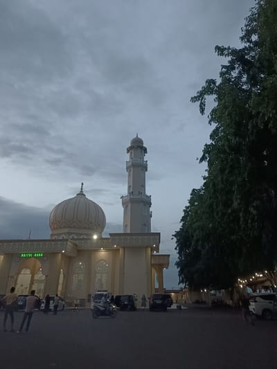 The image depicts the Baitul Huda Mosque in the city of Mee Tanjong Usi, Indonesia, during twilight. The mosque is a light-colored structure with a prominent dome and a tall minaret. The sky above is overcast with gray clouds. In front of the mosque, there is a paved area with several parked cars and a motorcycle. A few individuals are visible, some standing and walking. To the right of the mosque, a large tree with dark green foliage is prominent, and string lights are visible hanging from branches. The overall scene is dimly lit, characteristic of early evening or late afternoon before complete darkness.