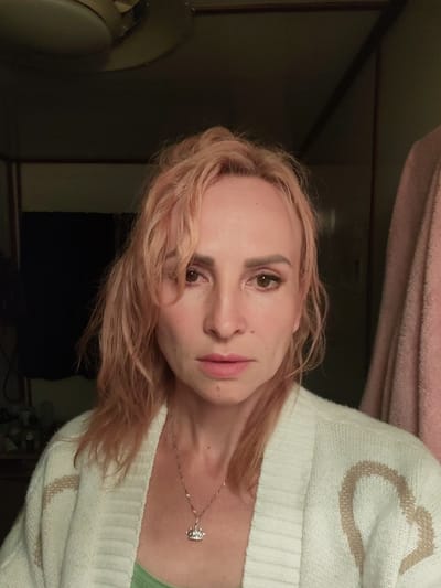 The image shows a close-up, front-facing, slightly overhead view of a person with blonde-pink hair. The person is wearing a cream-colored knitted cardigan with a tan heart pattern, and a light green undershirt. A delicate silver necklace with a crown-shaped pendant is visible on their neck. The person's gaze is directed towards the camera, and their expression is neutral. The background is dimly lit and appears to be an indoor environment, possibly a bedroom or dressing area, with hints of dark fabric, a ceiling fixture, and a pink textile hanging to the right. There is no discernible location within the city of Shalsky, Russia, indicated in the image.
