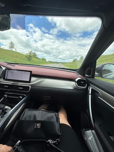 The image shows the interior of a car, likely a modern SUV, viewed from the passenger seat, looking forward through the windshield. The dashboard features a large touchscreen infotainment system, with a red and silver trim. To the left, a black handbag rests on the floor. The car is traveling on a paved road through a rural, grassy landscape with scattered trees. The sky is cloudy with patches of blue. The location is stated to be Kueda, Russia, though there are no specific visual cues in the image to confirm this. The scene captures a moment of travel on a road trip.