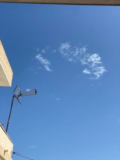 The image captures an upward perspective of a vibrant, clear blue sky over Rojales, Spain, illuminated by bright daylight. The vast expanse of blue is largely unobstructed, with a few scattered, wispy white clouds gracefully drifting in the upper and middle-left sections of the frame.
A prominent black and metallic television antenna dominates the mid-right portion of the composition. This antenna appears to be a combination of a Yagi-Uda array and a flat panel grid reflector, mounted on a diagonal pole that extends from the lower right corner upwards.
Architectural elements define the lower edges of the image. On the bottom-left, a sharp, angular edge of a light-colored, possibly beige or off-white, concrete or stucco structure is visible. Correspondingly, on the bottom-right, the edge of another similar light-colored building or roof is present. This structure includes a small, light-colored cylindrical pipe, likely a vent, and several black wires running across its surface. Another thin black cable extends into the frame from the far right.
No people are visible, and no specific activity or event is taking place; the image presents a serene, static view. The bright lighting and clear sky suggest a sunny day, possibly midday or early afternoon. The architectural style aligns with typical residential buildings in Mediterranean regions. There is no visible text in the image.