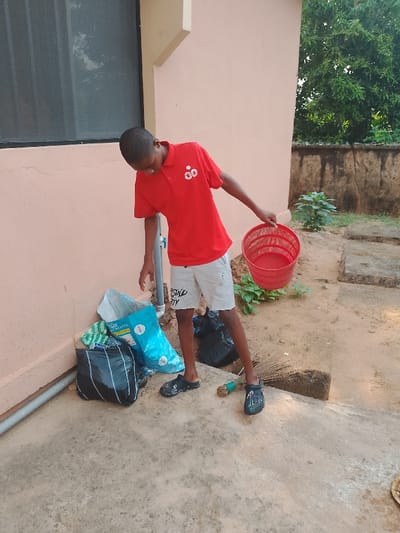 An outdoor scene in Ufuma, Nigeria, depicts a young male positioned on a concrete and dirt ground surface next to a light-peach colored building wall. The individual wears a red polo shirt with a white "GO" logo, light-colored shorts with "SOUND CITY" text on the right leg, and dark slip-on shoes. He holds a red, round, perforated plastic basket in his right hand, angled downwards, and is looking towards the ground near his feet. Against the building wall, to the left of the individual, is a pile of waste material: an open light-blue sack and at least two black plastic refuse bags. A white PVC pipe with a blue spigot is affixed to the wall next to this waste. A dark-framed window with a screen is visible on the upper left section of the wall. On the dirt ground near the individual's feet, a traditional broom constructed from dried plant material rests horizontally. The background features additional dirt ground with sparse green vegetation, a low, cracked concrete wall, and dense green trees and foliage under natural light.