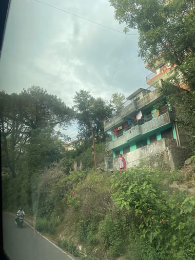 The image shows a multi-story building with green-painted balconies and grey concrete elements in what appears to be a hilly area. Clothes are hanging on the balconies to dry. The building is partially obscured by trees and other vegetation. The sky is overcast. The view is possibly taken from inside a vehicle, judging by the dashboard reflection. The location is Chelian, India. The image contains no visible people or apparent interactions. A road borders the treeline.