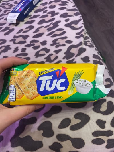 The image shows a hand holding a package of "TUC" crackers with a sour cream and onion flavor. The packaging is yellow and green with an image of the crackers and a bowl of sour cream with chives. The crackers themselves are thin and crispy, topped with herbs and spices. The background is a leopard-print fabric, possibly a blanket or bedspread. There is no information in the image or crops that provides any context about the city of Vaganovo, Russia.