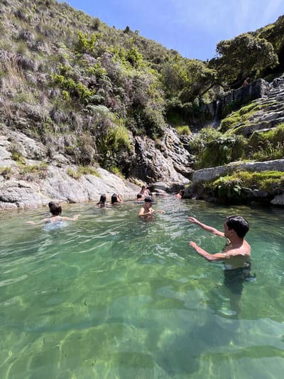 The image shows a natural pool of clear green water situated within a rocky, vegetated mountainous terrain. Several people are swimming and interacting in the water. In the foreground, a shirtless man with dark hair is partially submerged, extending his right arm forward. To his left, a person wearing a patterned swimsuit is visible from the back. Further into the pool, other individuals are seen swimming, with some with their heads above water. The background features steep, rocky inclines covered with lush green vegetation, grasses, and some trees. A stone staircase and a small waterfall are discernible on the right side of the frame, indicating a developed access point to the natural feature. The sky above is bright blue, suggesting a sunny day. The location is identified as a natural pool in Mucurubá, Venezuela.