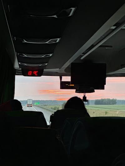 The image is taken from inside a bus, looking forward through the windshield. The sky outside is a vibrant orange and pink, suggesting either sunrise or sunset. The road ahead shows a truck and a green sign. Inside the bus, a digital display shows "13 °C". Several passengers are visible in silhouette in the foreground, seated on the bus. The interior of the bus features overhead compartments and a screen mounted to the ceiling. The scene captures a moment of travel, with the outside environment contrasting with the interior. No specific landmarks or identifying features of Ligota Polska, Poland are visible within the frame.