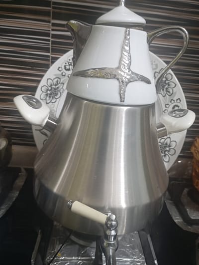 The image depicts a beverage dispenser in an indoor setting within the city of Nasiriyah, Iraq, likely a kitchen or buffet area. The central object is a two-part, samovar-style dispenser. The upper section is white, possibly ceramic or enamel, with a conical body adorned with prominent silver-colored, textured, cross-shaped embellishments. It features a silver-colored spout on the left, a curved silver handle on the right, and a white lid topped with a silver finial. The lower section is constructed from brushed stainless steel, presenting a wider, inverted conical form. A chrome-plated tap with an off-white handle extends from the lower front of the stainless steel base. Behind the dispenser, a white plate with intricate black and grey floral patterns is partially visible. The background wall is tiled with alternating horizontal stripes of dark brown and light beige. The dispenser rests on a dark surface, partially covered by crumpled aluminum foil. Peripheral elements include a dark, indistinct object on the far left and a textured, possibly wicker, item on the far right. No individuals, actions, or interactions are visible in the static scene.