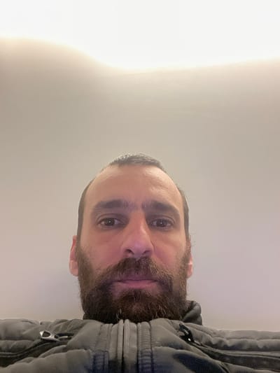 The scene is located in Etterbeek, Belgium. A single adult male is depicted in a close-up, low-angle photograph, occupying the lower half of the frame. The subject has a medium complexion, dark eyes, dark hair, and a full dark beard and mustache. His hair on the scalp is dark and shows a receding hairline. He is wearing a dark grey, likely padded or quilted, zip-up jacket with a visible central zipper and a black zipper pull or clip on the left side of the chest. The subject's gaze is directed forward, specifically into the camera lens. The background consists of a plain, light-colored wall or ceiling, with a brighter, less distinct area in the upper portion, suggesting an overhead light source or bright ceiling. The environment appears to be indoors. No actions or interactions beyond the subject's direct gaze are evident.