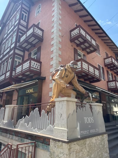 This vertically oriented image shows the exterior of a building with ornate balconies and windows. A statue of a person in a tan jumpsuit, goggles, and backpack is depicted climbing up the side of the building. The statue is a prominent subject, positioned in the center of the frame. Below the statue, a sign reads "TOD'S FOR FRANZ KRALER" and another sign for "LOEWE" is visible on a shopfront. The building appears to be a commercial establishment, possibly a luxury store, given the brands displayed. The architecture suggests a European setting, and the text "119 CORSO ITALIA" further confirms an Italian location, specifically Cortina d'Ampezzo as hinted. The sky above is blue with some wispy clouds, indicating daytime and fair weather. The building's facade is a warm pinkish-red, contrasting with the dark brown of the balconies and window frames, and the white decorative elements on some of the balconies. The overall scene is likely from a street in a town, with a focus on the building's architecture and a striking artistic installation.