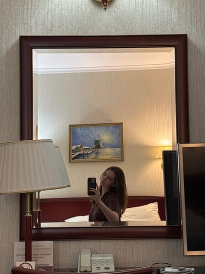 The image appears to be a reflection in a mirror within a hotel room. A young woman is visible in the mirror, taking a selfie with her phone. She has long, brown hair and is wearing a brown shirt. Behind her, a painting of a windmill and houses by the water is hung on the wall. A bedside lamp with a pleated shade is also visible. In the foreground, a phone and some charging cables are on a table. The room has textured wallpaper and a dark wooden mirror frame. The provided location, Журавлево (кемеровский), Russia, suggests this scene is set in a hotel room in that specific region.