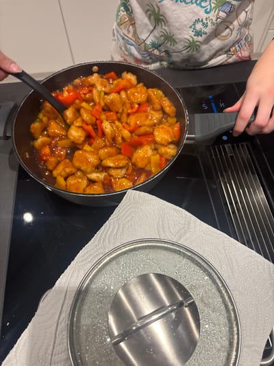 A frying pan containing a mix of diced chicken and red and yellow bell peppers in a sauce sits on a black stovetop. A black spatula is in the pan mixing the ingredients. A nearby pan lid sits on a white paper towel. A person's hand is shown adjusting the stovetop controls, while part of a person wearing a white shirt with palm trees is also in the shot.