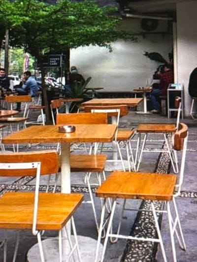 The image depicts an outdoor seating area, possibly a cafe, with several wooden tables and chairs. In the background, a man wearing a mask is visible, and further back, two individuals are seated at a table. Trees and foliage are present, suggesting an urban outdoor setting. The ground is paved with a textured material. There is an ashtray on one of the tables. The overall scene is casual and relaxed, with the focus on the dining and seating arrangements. The scene appears to be in Babahjurong, Indonesia, given the contextual information provided.