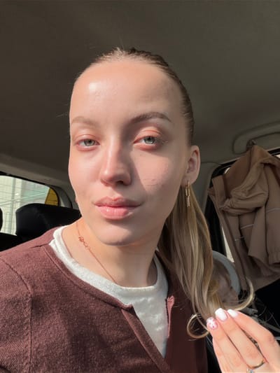 The image displays a close-up portrait of an adult female individual seated inside a vehicle. The subject has fair skin, light brown hair pulled back into a ponytail, and green-blue eyes. She is wearing a white top beneath a dark brown v-neck cardigan. Her lips appear to have a glossy finish. Accessories include gold-toned chain-link earrings on her right ear (viewer's left), a delicate gold-toned necklace with a small heart-shaped charm, and a ring with a light blue stone on her left ring finger (viewer's right). Her left hand is partially raised, displaying manicured nails painted white with small pink floral or dot patterns on the ring and pinky fingers. Bright natural light illuminates the scene from the left, casting shadows across the subject's face. The background reveals an automobile interior with a dark headliner, a dark car seat visible to the left, and a light-colored jacket hanging on a hook on the right side of the vehicle's cabin. A blurred exterior view is visible through the window on the left. The specific location within the city of Bezborodovo, Russia, cannot be determined from the visual information provided.