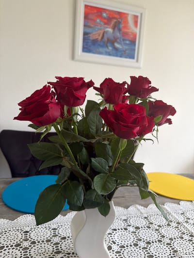A vase of red roses sits on a table next to a crocheted lace tablecloth and a colorful wall decoration. The vase appears to be white with yellow and blue accents. A framed artwork hangs on the wall in the background. While the image doesn't explicitly show the location is Rapperswil-Jona, Switzerland, I cannot confirm that detail from the image itself.