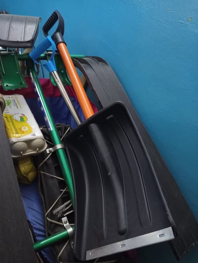 In Novotroitsk, Russia, this image depicts a collection of winter tools and supplies, likely stored in a shed or garage. A large black plastic snow shovel with a metal edge is prominently featured, leaning against a dark wooden surface. To its left, a bright orange shovel handle with a black grip is visible, along with a blue D-shaped handle attached to a green pole. Several other green poles, likely belonging to other snow removal tools, are also present. A carton of eggs, showing Cyrillic text and a cartoon chicken, rests on a blue fabric surface, suggesting this storage area may also be used for household goods. The background is a textured, bright blue wall. The scene is lit by an overhead light source, casting subtle shadows and highlighting the textures of the objects.