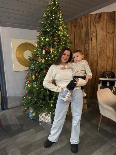 The scene is observed indoors, likely within a residence or establishment in Kalinovo, Russia. A woman stands centrally, holding a young child. The woman has long dark hair and wears a cream-colored, long-sleeved, ribbed sweater, light blue wide-leg jeans, and black chunky-soled shoes. Her left hand rests on her hip, and her right arm supports the child. She is looking towards the viewer with a slight smile. The child, a toddler, is dressed in a light-colored striped hooded long-sleeved top, dark pants, and metallic silver boots with visible blue and yellow socks. The child's head is turned to the left, away from the camera.

Behind the individuals, a tall, green artificial Christmas tree is heavily decorated with numerous warm white lights, red and gold spherical ornaments, teardrop-shaped ornaments, and various star and snowflake decorations. Several wrapped gift boxes, featuring patterns such as blue and white, are placed at the base of the tree.

The background consists of two distinct wall sections. To the left of the tree, a plain light grey wall displays a large white square canvas with a central golden concentric circle design. To the right of the tree, a wall is constructed from vertical, irregularly shaped planks of naturally finished wood. In the far right background, a beige high chair with a black tray is visible next to a light-colored armchair with wooden legs. The floor throughout the visible area is dark grey with a herringbone pattern.