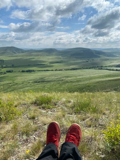 A person's legs, clad in black trousers and red sneakers, are visible in the foreground of this image. They are seated on a grassy hill overlooking a vast, rolling landscape of green hills and valleys under a partly cloudy sky. The terrain appears to be rural, with sparse trees dotting the landscape in the middle ground. The location is identified as Andreevka, Russia.