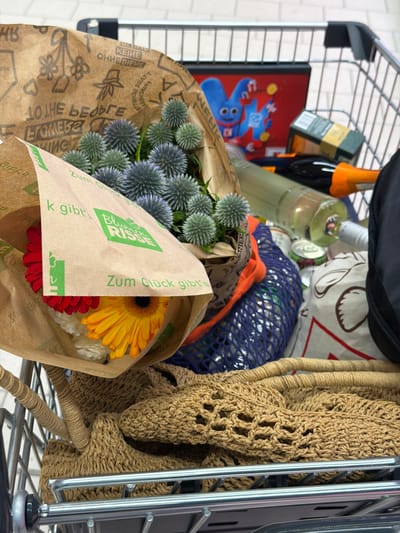 The image shows the interior of a shopping cart filled with various items. In the foreground, a bouquet of blue thistles and gerbera daisies, one red and one yellow, are partially visible, wrapped in brown paper with green text that includes "Blumen Risse". Next to the flowers, a blue mesh bag is visible, containing what appear to be cans. Further back, a bottle of white wine with a green label and an orange cap is present, alongside another bottle of wine and a red box with a cartoon character. A black bag is also visible in the upper right corner. The shopping cart itself is made of metal and appears to be in a store environment, as suggested by the tiled background. The overall impression is that of a shopping trip, possibly for groceries and flowers. The presence of the "Blumen Risse" branding suggests a location where this flower shop operates. However, there is no explicit information in the image to pinpoint the city of Witten, Germany.