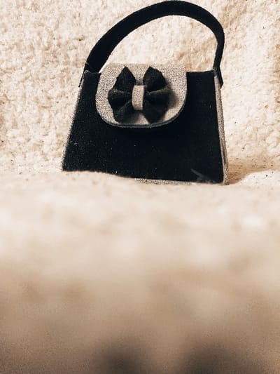 A small, black handbag with a structured body, a single handle, and a semi-circular light grey flap is positioned upright in the center of the frame. A black bow is affixed centrally to the grey flap. The bag appears to be made of a matte, possibly suede or velvet-like material. The item rests on a light-colored, highly textured surface, consistent with a shag carpet or thick fleece fabric, which also forms the background. The immediate foreground is intentionally blurred, presenting as a lighter, amorphous area. The image is rendered with a desaturated, sepia-toned effect. No individuals, actions, or interactions are present. The image contains no visual information that indicates its location in the city of Harasta, Syria.