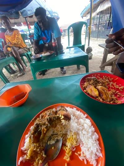 The image depicts an outdoor dining establishment in Naze, Nigeria, operating under a blue and white striped canopy. In the immediate foreground, two red plates are positioned on a green plastic table. The plate on the left holds white rice accompanied by a dark green stew containing chicken pieces, with a metal spoon resting within the food. An orange plastic bowl containing a red dispenser is situated to the left of this plate. The partially visible plate on the right contains reddish rice, fried plantains, and dark protein items; a dark-skinned hand holding a white utensil is poised over it, with a dark-colored mobile phone visible nearby. In the mid-ground, a dark-skinned man wearing a light blue graphic t-shirt and flip-flops is seated at an adjacent green plastic table, leaning over a bowl of food. A glass Coca-Cola bottle is placed next to his meal. To his left, a dark-skinned woman in a yellow off-shoulder top and sandals sits on a green plastic chair, looking forward. The ground beneath the tables is exposed earth, and the background reveals further green plastic chairs and distant buildings featuring metal security bars on windows, indicative of a street-side setting.