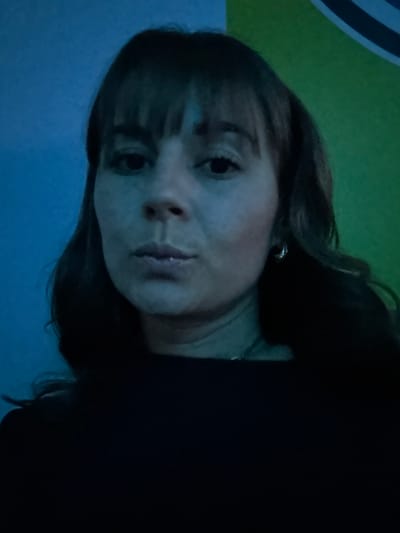 A woman with bangs and dark hair faces the camera. She wears a dark garment, possibly a sweater or shirt. Her skin appears pale, likely due to lighting conditions. She has small, subtle earrings. The background features a mix of blue and green hues, suggesting a wall or other surface with varying colors or lighting. The image is slightly tilted, giving a casual, slightly off-kilter impression.