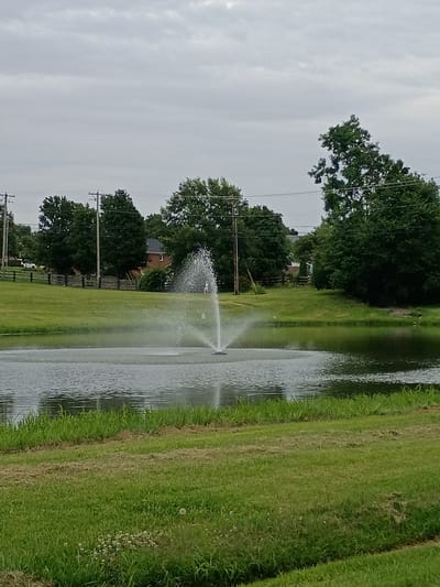 This image depicts an outdoor scene with a pond and a fountain in the foreground. The pond's surface is disturbed by the fountain's spray, creating a misty effect. The surrounding area is covered in green grass, with some areas appearing more manicured than others, suggesting a park or well-kept residential area. In the background, there are trees, power lines, and a house with a red brick exterior. A wooden fence runs along a grassy embankment. Two small figures, possibly people, are visible on the grass near the fountain. The sky is overcast with a grey, cloudy appearance. The location is identified as Wilmore, United States of America, based on the context provided.
