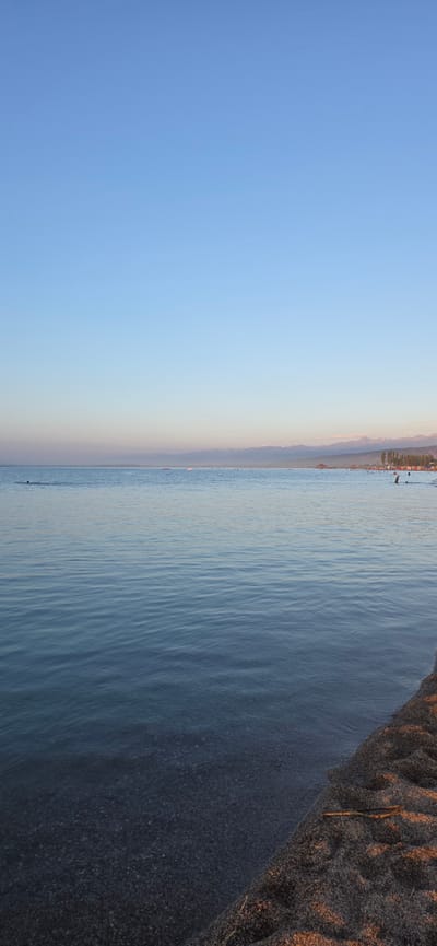 The image depicts a serene beach scene at the city of Boz Beshik, Kyrgyzstan. The foreground features a sandy shore with a few small sticks scattered on it. The water is calm with gentle ripples, and the pebbles beneath the surface are visible. In the distance, a thin line of trees and structures can be seen along the coastline, with a mountain range forming the backdrop under a clear, pale blue sky. Several people are present in the water, some swimming and others appearing to be relaxing, while a few more are visible on the distant shore. The overall atmosphere is peaceful, suggesting a tranquil day at the beach.
