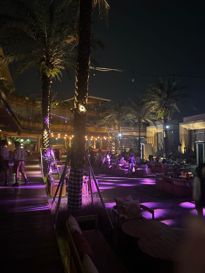 The image depicts a nighttime scene in Kosambi Barat, Indonesia. The setting appears to be an outdoor dining area or patio, illuminated by string lights and purple spotlights. Tables and chairs are arranged, and several people are seated and interacting, suggesting a social atmosphere. Palm trees are visible, adding to the ambiance. The environment is urban, with buildings visible in the background. One person is walking along the edge of the patio.
