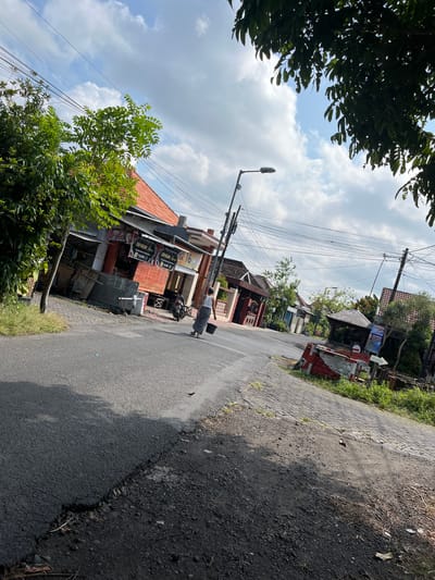 The image depicts a street scene in the city of Jati Alun Alun, East Java, under a partly cloudy sky. The street is paved with asphalt, and a cobblestone path runs alongside it. A person wearing a white tank top and checkered pants is walking away from the camera, carrying a black bucket in their right hand. To the left of the street, there are several buildings with orange tile roofs, some appearing to be shops with signage. A tree with green foliage is situated near the buildings. A lamppost stands on the side of the road, and numerous power lines crisscross overhead. In the background, more buildings and vegetation are visible, suggesting a residential or commercial area. A motorcycle is parked near one of the buildings.