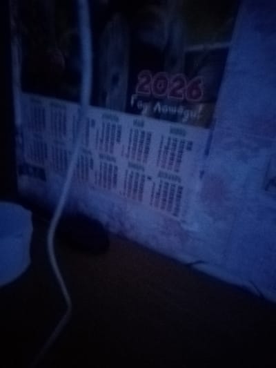 The image shows a close-up of a calendar with the year "2026" prominently displayed in pink lettering. Below the year, there is text in Russian that appears to read "Год Лошади" which translates to "Year of the Horse". The calendar pages are visible with columns and rows of text, likely days of the week and dates. The overall lighting is dim and blue-toned, suggesting it was taken in low light conditions. The calendar is positioned on a surface that appears to be a desk or table, made of wood. A white cord or cable is visible running diagonally across the calendar and into the lower left portion of the frame, where a soft, out-of-focus object, possibly a cushion or cloth, is present. The background is dark and blurred, with hints of other objects that are not clearly identifiable. The image's quality is grainy, indicating it was likely taken with a mobile device in a low-light environment. There are no people visible in the image, and the focus is on the calendar itself. The location of Советская Хакасия, Russia is not discernible from the image content itself.