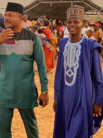 This vertical image depicts an outdoor gathering in Agunu, Nigeria, likely under a temporary canopy or shelter. In the foreground, two men stand amidst a crowd.
The man positioned higher in the frame is smiling broadly and looking directly at the camera. He wears a flowing royal blue traditional garment, possibly an agbada, featuring intricate white embroidery on the chest, and a multi-patterned cap.
Below him, another man looks downwards and slightly away from the camera, with a contemplative expression. He is dressed in a dark green two-piece outfit, possibly a traditional or 'senator style' suit, with a geometric pattern in light grey and black on the upper chest and shoulders. He wears a black cap, a gold cross pendant, and a prominent gold wristwatch on his left wrist. He is holding a dark-colored mobile phone in his right hand.
The background shows a large group of people, including men, women, and children, suggesting a community event, political rally, or religious gathering. They are dressed in a variety of casual and traditional attire. The ground is a dry, sandy brown. The bright illumination indicates it is daytime. There is no visible text in the image.