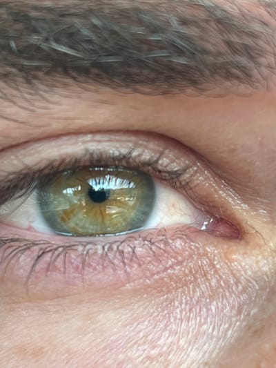 The image shows a close-up of a person's right eye. The eye is open and looking towards the right. The iris is green with brown flecks, and the pupil is black. There are several eyelashes visible on the top and bottom eyelids. The skin around the eye is fair and there is a slight red blood vessel visible on the lower eyelid. The eyebrow is visible above the eye. The location of the image is near Kiryat Ye'arim, Israel, based on the coordinates provided.