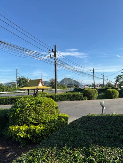 The image shows a street scene in Ban Ko Pru, Thailand, with utility poles and lines on the left side of the frame extending into the sky. A small, yellow-roofed structure sits along the roadside, partially obscured by lush green shrubbery. A narrow paved road runs parallel to the shrubbery, with more green plants on the right side of the frame. In the background, a mountain is visible under a clear blue sky with scattered white clouds. A KFC sign can be seen in the distance along the roadside.