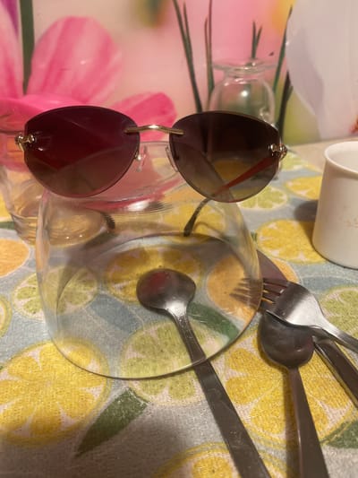 A pair of rimless sunglasses with dark lenses and gold-colored trim rests on the rim of a clear glass bowl. The bowl is placed on a tablecloth with a pattern of yellow and green citrus slices. To the right of the bowl, a white mug is visible. Several pieces of silverware, including spoons and a fork, are scattered around the bowl. In the background, out of focus, are large pink floral elements and a small glass vase with green stems. The image appears to be an indoor still life. The location is not specified in the image.
