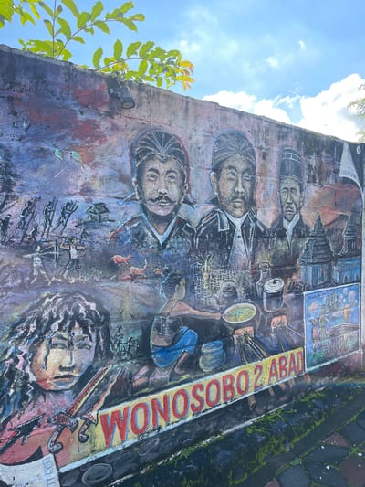 A mural covers an exterior wall in Sinduagung, Indonesia. It depicts four Indonesian men in traditional attire and headwear in an overlapping vertical stack. Above them, a building and scenery are depicted. The word "WONOSOBO 2 ABAI" in red letters is painted on a yellow stripe. Below the men, stick figures march forward with muskets. A woman's face lies on the bottom part of the wall next to a dagger. A tree branch juts out from the left side of the wall. A light fixture is attached to the left side of the wall. Paved tiles lie beside the wall. The sky with clouds is visible in the top left of the image.
