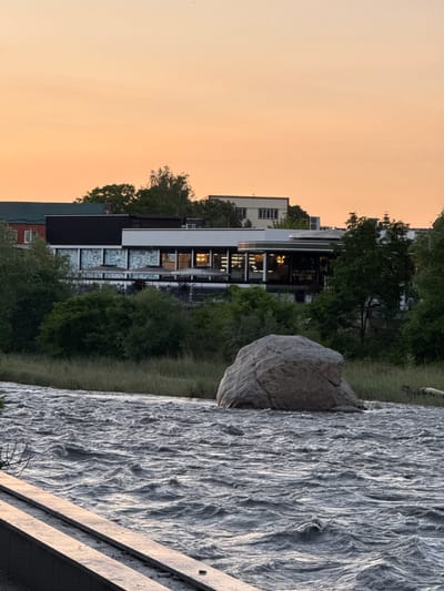The image depicts a river flowing rapidly in the foreground, with white caps and choppy waves indicating a strong current. A large, smooth boulder rests on the riverbank on the right side of the frame, partially submerged in the water. The riverbank is covered in tall, dry grass. Behind the riverbank, a modern building complex is visible, featuring a white facade with dark accents and large windows. Several people are seated at tables on an outdoor patio area in front of the building. Above the building, trees and other structures are visible, including a taller, white building further in the background. The sky is a gradient of soft orange and yellow hues, suggesting either sunrise or sunset. The scene appears to be a waterfront area in a city, possibly Askhazan, Russia, with a focus on the natural elements of the river and the man-made structures.