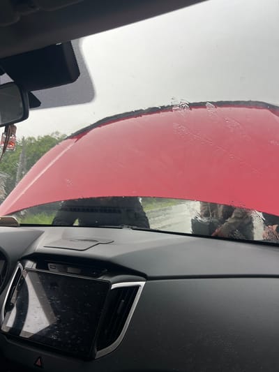 The image provides a vertical view from inside a vehicle, looking out through a wet windshield. Water droplets are visible across the windshield and on interior surfaces, suggesting rain or recent rainfall.
The dominant external subject is a large, featureless red surface, likely the side of a truck, train car, or another large object, seen through the driver's side of the windshield. This red surface appears textured or painted and is also wet. A distinct dark, vertical border separates this red mass from a lighter background. Faint reflections or views of green foliage, possibly trees, are visible in the background, indicating an outdoor setting.
Inside the vehicle, the black, textured dashboard is prominent. On the right side, a large infotainment screen, covered in water droplets and smudges, is clearly visible. This screen has a black bezel and shows indistinct touch icons along its bottom edge. Adjacent to the screen is an air vent with a silver trim. A red triangular hazard light button is partially visible near the bottom right of the screen. On the left side of the windshield, parts of the rearview mirror assembly are visible, including a black sensor housing with a dotted pattern on the glass. A small, clear plastic object with a red switch is attached to the mirror area.
The scene depicts a stationary vehicle, possibly stopped during or after a period of rain, next to a large red object. The time of day appears to be daytime, with overcast conditions suggested by the diffused light. No people are visible, and no discernible text is present in the image.