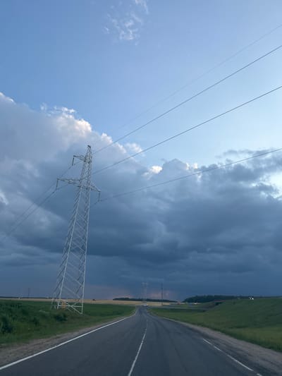 The image shows a road leading into the distance under a dramatic sky with dark storm clouds. A tall, metal electricity pylon stands to the left of the road, with power lines stretching across the frame. The landscape consists of green fields and some trees in the distance. The road appears to be an asphalt highway with white lane markings. The lighting suggests it is either early morning or late afternoon, with the sun attempting to break through the clouds, creating a muted, atmospheric effect. The scene is devoid of any people but shows some vehicles in the distance on the road and a few structures that appear to be agricultural or industrial. The location is identified as Zhukhovichy Village Council, Belarus.